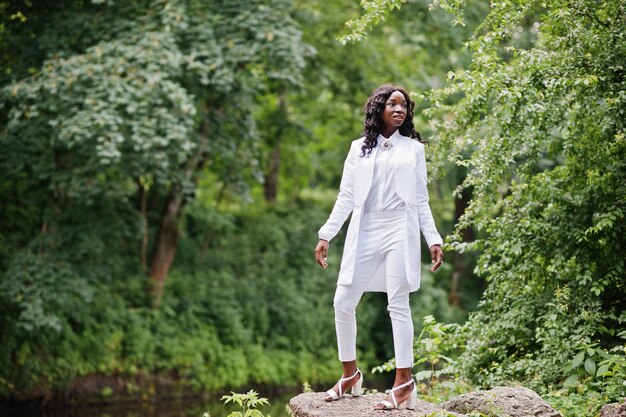 Elegante chica afroamericana negra posando en piedra en el río de fondo del parque