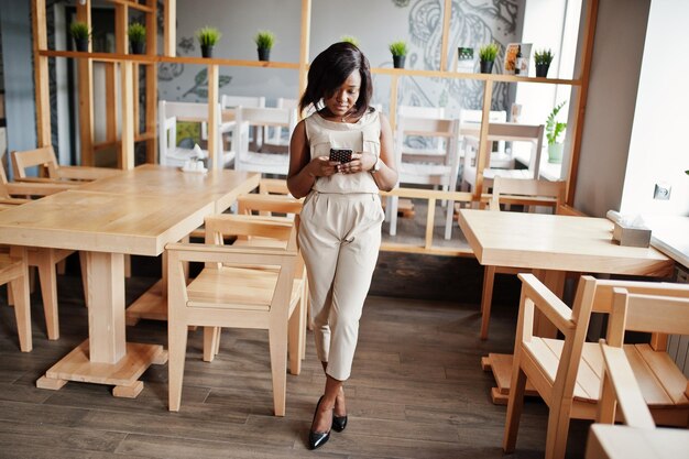 Elegante chica afroamericana posó en un café moderno y mirando el teléfono móvil