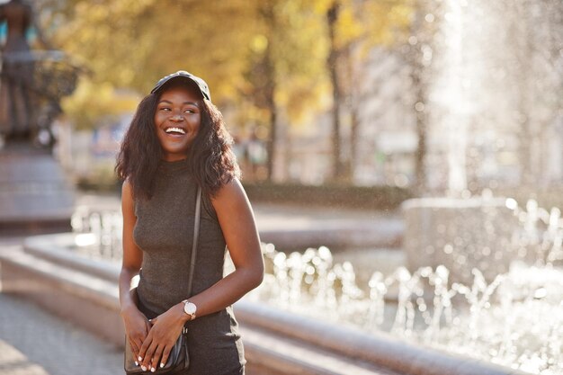 Elegante chica afroamericana en túnica gris bandolera y gorra posada en el soleado día de otoño contra fuentes África mujer modelo