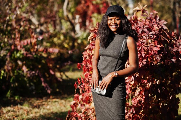 Elegante chica afroamericana en túnica gris bandolera y gorra posada en el soleado día de otoño contra hojas rojas África modelo mujer