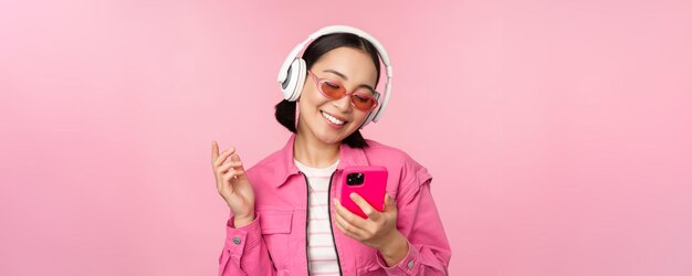 Elegante chica asiática bailando con smartphone escuchando música en auriculares en la aplicación de teléfono móvil sonriendo y riendo posando sobre fondo rosa