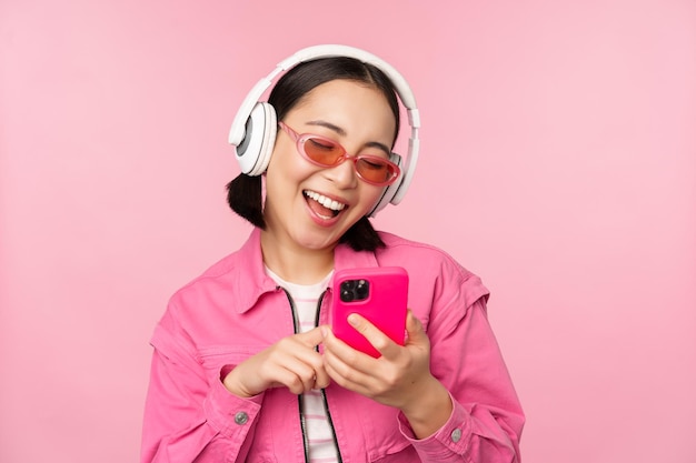 Elegante chica asiática bailando con smartphone escuchando música en auriculares en la aplicación de teléfono móvil sonriendo y riendo posando sobre fondo rosa