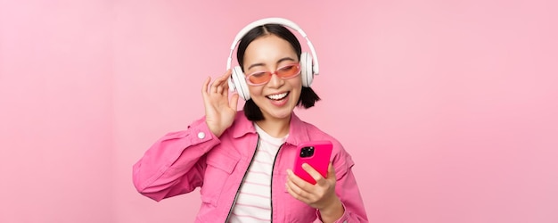 Elegante chica asiática bailando con smartphone escuchando música en auriculares en la aplicación de teléfono móvil sonriendo y riendo posando sobre fondo rosa