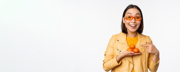 Elegante chica asiática feliz con gafas de sol sosteniendo mandarinas y sonriendo posando sobre fondo blanco Copiar espacio