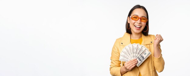 Elegante chica asiática sonriente sosteniendo dinero en efectivo mostrando dólares y celebrando de pie sobre ba blanca