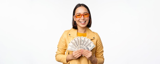 Elegante chica asiática sonriente sosteniendo dinero en efectivo mostrando dólares y celebrando de pie sobre fondo blanco.