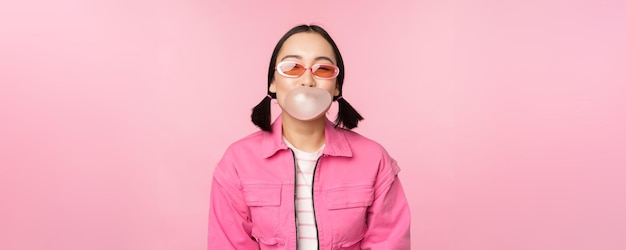 Foto gratuita elegante chica asiática soplando chicle con gafas de sol posando sobre fondo rosa copiar espacio