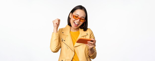 Elegante chica coreana con gafas de sol jugando videojuegos móviles riendo y sonriendo mientras usa un teléfono inteligente de pie sobre fondo blanco.