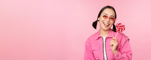 Elegante chica coreana lamiendo lolipop comiendo dulces y sonriendo de pie en gafas de sol contra bac rosa