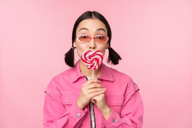 Elegante chica coreana lamiendo lolipop comiendo dulces y sonriendo de pie en gafas de sol contra el fondo rosa