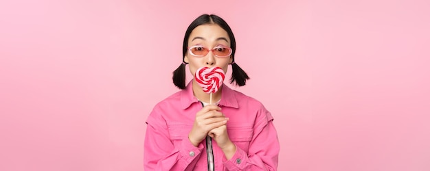 Elegante chica coreana lamiendo lolipop comiendo dulces y sonriendo de pie en gafas de sol contra el fondo rosa