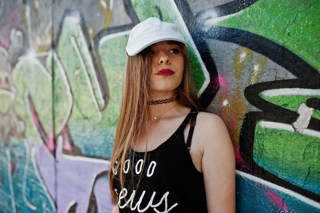 Elegante chica hipster casual con gorra y jeans contra una gran pared de graffiti