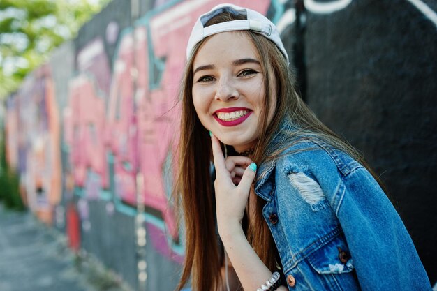 Elegante chica hipster casual con gorra y jeans usa música para escuchar desde auriculares de teléfono móvil contra una gran pared de graffiti con bomba