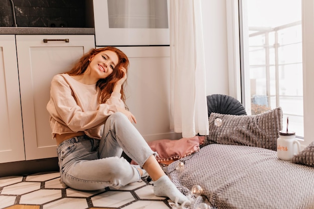 Elegante chica jengibre relajándose en casa Mujer riendo pelirroja en jeans sentada en el suelo