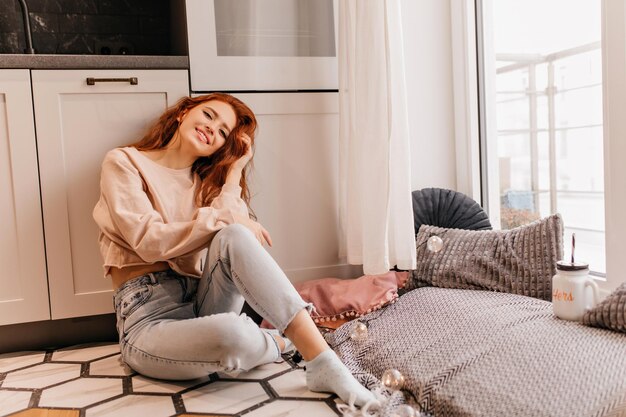 Elegante chica jengibre relajándose en casa Mujer riendo pelirroja en jeans sentada en el suelo