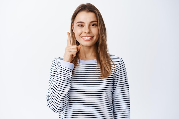 Elegante chica moderna con cabello rubio y piercing, apuntando con el dedo hacia arriba y sonriendo, teniendo una idea o sugerencia, vistiendo ropa casual, blanco