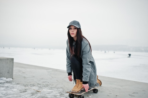 Elegante chica morena con gorra gris estilo casual street con patineta en el día de invierno