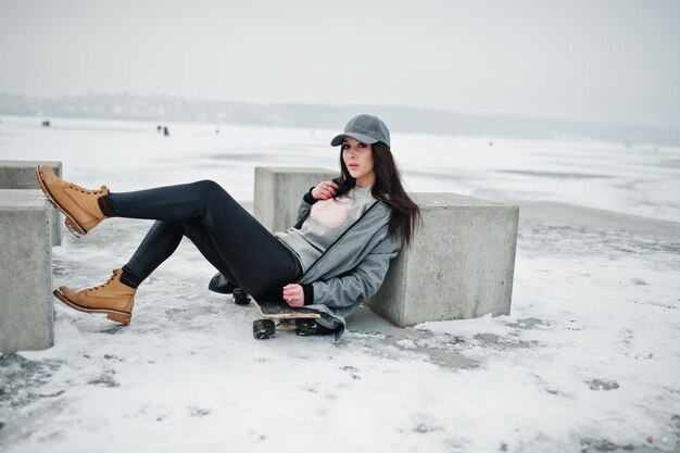 Elegante chica morena con gorra gris estilo casual street con patineta en el día de invierno