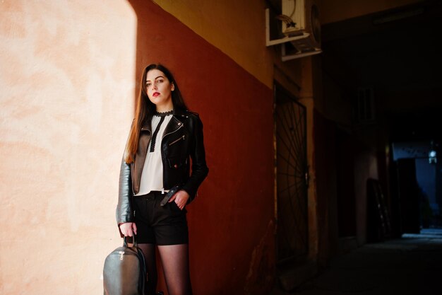 Elegante chica morena vestida con chaqueta de cuero y pantalones cortos con mochila contra la pared naranja en las sombras