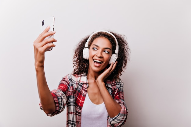 Elegante chica negra escuchando música mientras toma una foto de sí misma. Mujer entusiasta con teléfono para selfie.