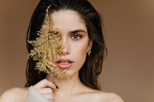 Elegante chica pálida con aretes dorados que expresa asombro. Toma de primer plano de dama morena sensual con planta.