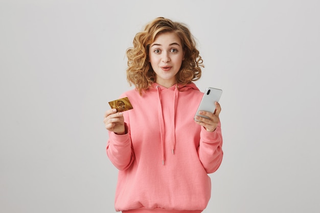 Elegante chica de pelo rizado de compras en línea, con tarjeta de crédito y teléfono inteligente