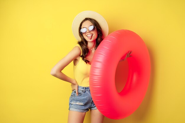 Elegante chica de playa con anillo de natación, riendo y sonriendo en vacaciones de verano, viaje por mar, de pie feliz contra el fondo amarillo.