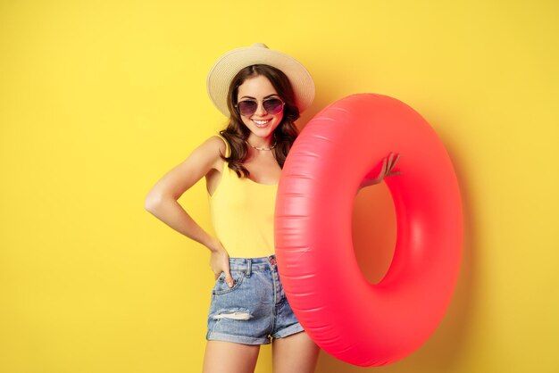 Elegante chica de playa con anillo de natación, riendo y sonriendo en vacaciones de verano, viaje por mar, de pie feliz contra el fondo amarillo.