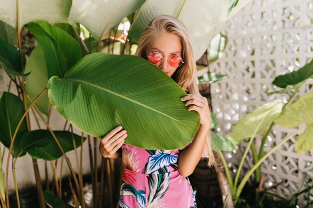 Elegante chica rubia lleva gafas de sol elegantes que se esconden detrás de la hoja grande.