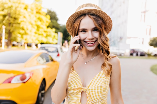 Elegante chica rubia con sombrero vintage hablando por teléfono en fin de semana de verano. Modelo femenino blanco lindo en traje amarillo que disfruta del paseo matutino.