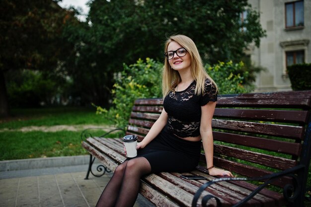 Elegante chica rubia vestida de negro posando en las calles de la ciudad sentada en un banco con una taza de café en las manos