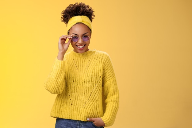 Elegante y coqueta mujer afroamericana moderno peinado afro en suéter diadema tocando gafas de sol s ...