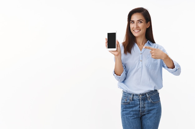 Elegante dama adulta alegre, guapa, complacida, promover el teléfono inteligente, sostener el teléfono móvil, señalar la pantalla del dispositivo, recomendar el uso de la aplicación, mostrar la visualización de las fotos de las vacaciones de verano, sonreír felizmente