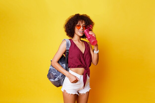 Elegante deporte mujer negra de pie sobre amarillo y sosteniendo una botella de agua rosa Vistiendo elegante ropa de verano y mochila.