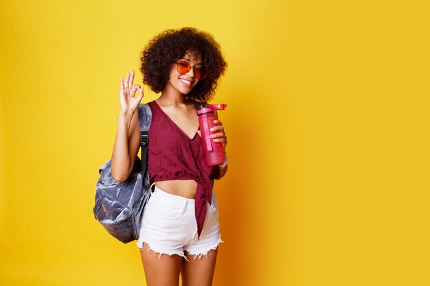 Elegante deporte mujer negra de pie sobre amarillo y sosteniendo una botella de agua rosa Vistiendo elegante ropa de verano y mochila.