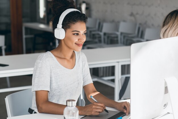 Elegante diseñadora web africana haciendo su trabajo en la oficina con una sonrisa. Atractiva mujer negra en auriculares blancos trabajando en call-center, sentado junto a la computadora.