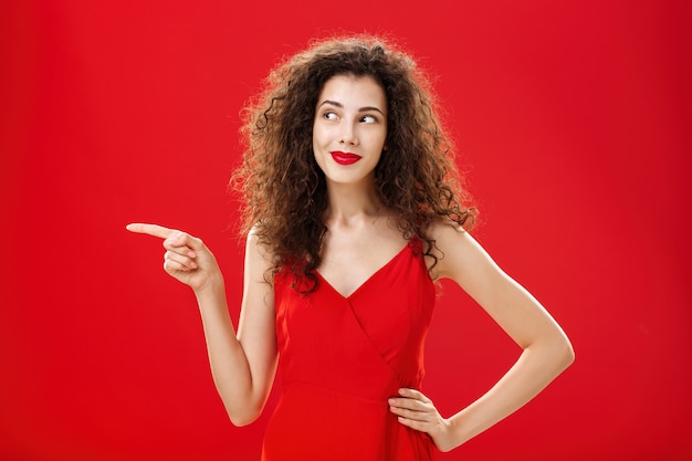 Elegante y elegante mujer encantadora curiosa con peinado rizado en lápiz labial rojo y vestido de moda sosteniendo la mano en la cadera apuntando y mirando a la izquierda encantada e interesada sobre el fondo del estudio.