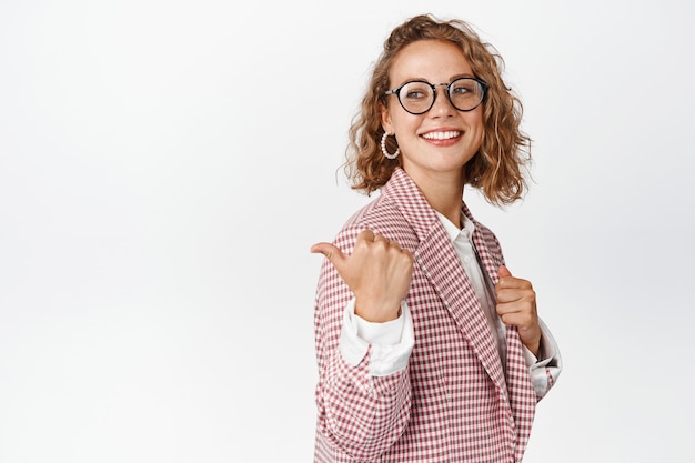 Elegante empresaria con gafas y traje, sonriendo y mostrando algo, señalando con el dedo el logotipo de la empresa, espacio de copia del lado izquierdo en blanco.