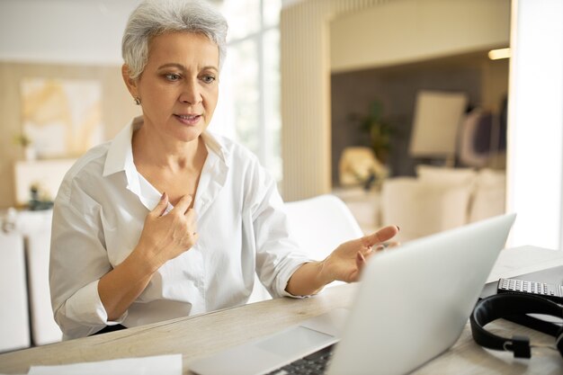 Elegante empresaria madura con corte de pelo corto sentado frente a la computadora portátil, mirando la pantalla con la boca abierta como si dijera algo