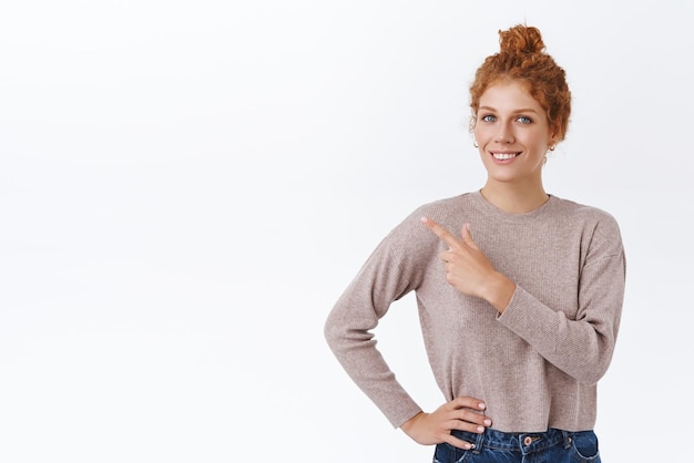 Elegante empresaria pelirroja guapa y asertiva con cabello rizado en un elegante suéter que apunta a la esquina superior izquierda, sostenga la mano en la cintura, cámara sonriente, anuncie la promoción del producto