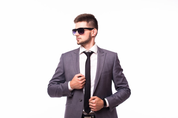 Elegante y encantador. Retrato de joven guapo en ropa formal y gafas de sol ajustando su corbata mientras está de pie contra el fondo gris