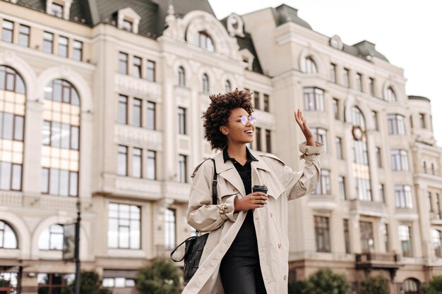 Elegante y encantadora dama morena de piel oscura con anteojos, gabardina beige y vestido negro, sonríe, saluda con la mano y sostiene una taza de café al aire libre