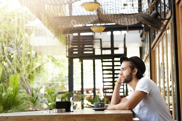 Elegante estudiante de barba caucásica en sombreros bebiendo capuchino solo en la moderna cafetería al aire libre en un día soleado, esperando que su novia se una a él