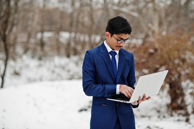 Elegante estudiante indio con traje y anteojos posó en el día de invierno al aire libre con una computadora portátil en las manos