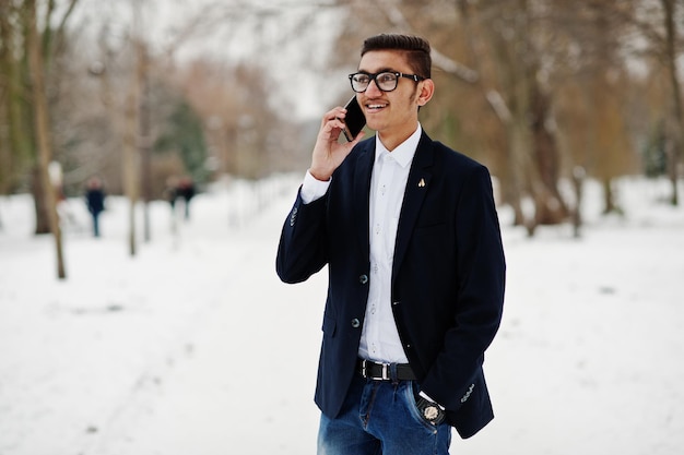 Elegante estudiante indio con traje y anteojos posó en el día de invierno al aire libre y hablando por teléfono móvil