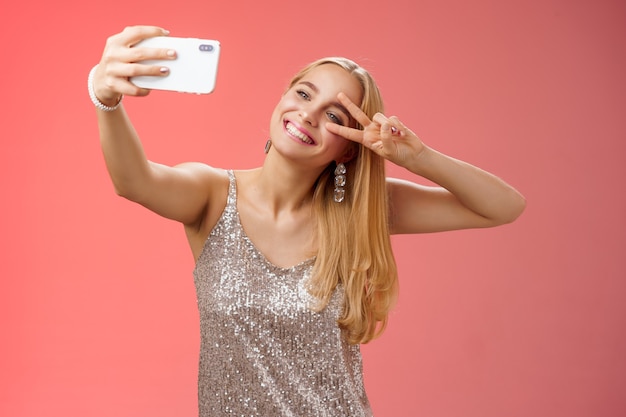 Elegante glamour fabulosa joven rubia en vestido plateado brillante inclinando la cabeza sin preocupaciones mostrar gesto de paz signo de victoria extender el brazo sosteniendo el teléfono inteligente tomando selfie grabación de video en línea