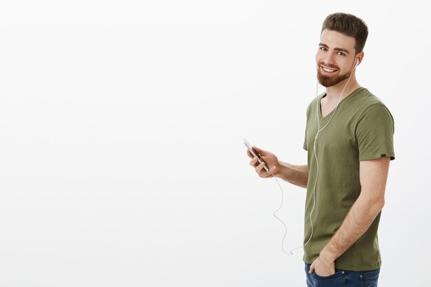 Elegante, guapo, descarado, hombre con barba en camiseta de pie en profily girando, sonriendo, sosteniendo un teléfono inteligente y usando auriculares mientras escucha música sobre una pared blanca