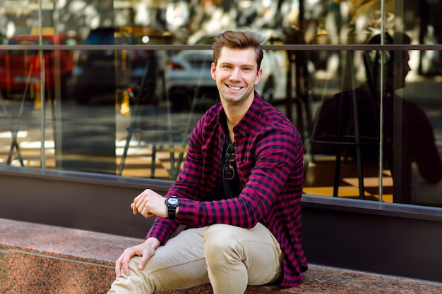 Elegante y guapo joven empresario sentado en la calle, increíble sonrisa, cabello castaño y ojos, vestido con camisa a cuadros hipster y pantalones beige, gafas de sol y relojes, posando cerca del restaurante.