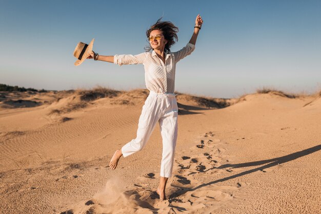 Elegante hermosa mujer alegre sonriente corriendo feliz en la arena del desierto vestida con pantalones blancos y blusa con sombrero de paja en la puesta del sol