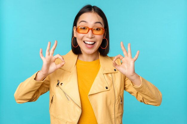 Elegante hermosa mujer asiática con gafas de sol sonriendo asombrada mostrando bien bien firmar recomendando algo apoya una excelente elección de pie sobre fondo azul